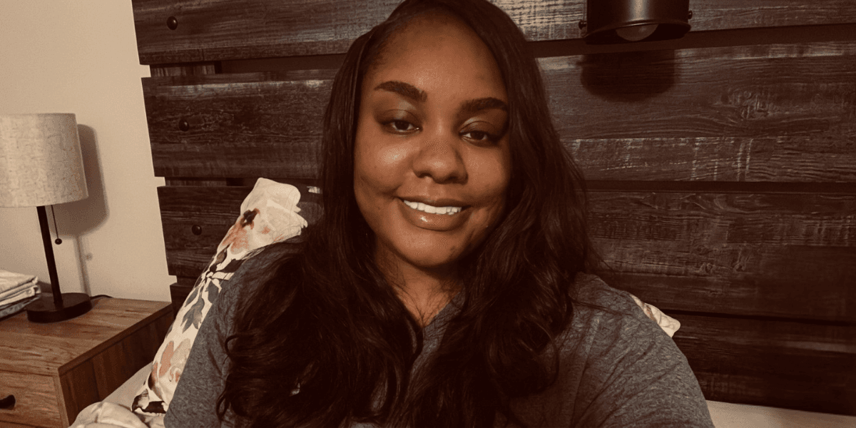 A woman with long hair smiles in a cozy bedroom setting.