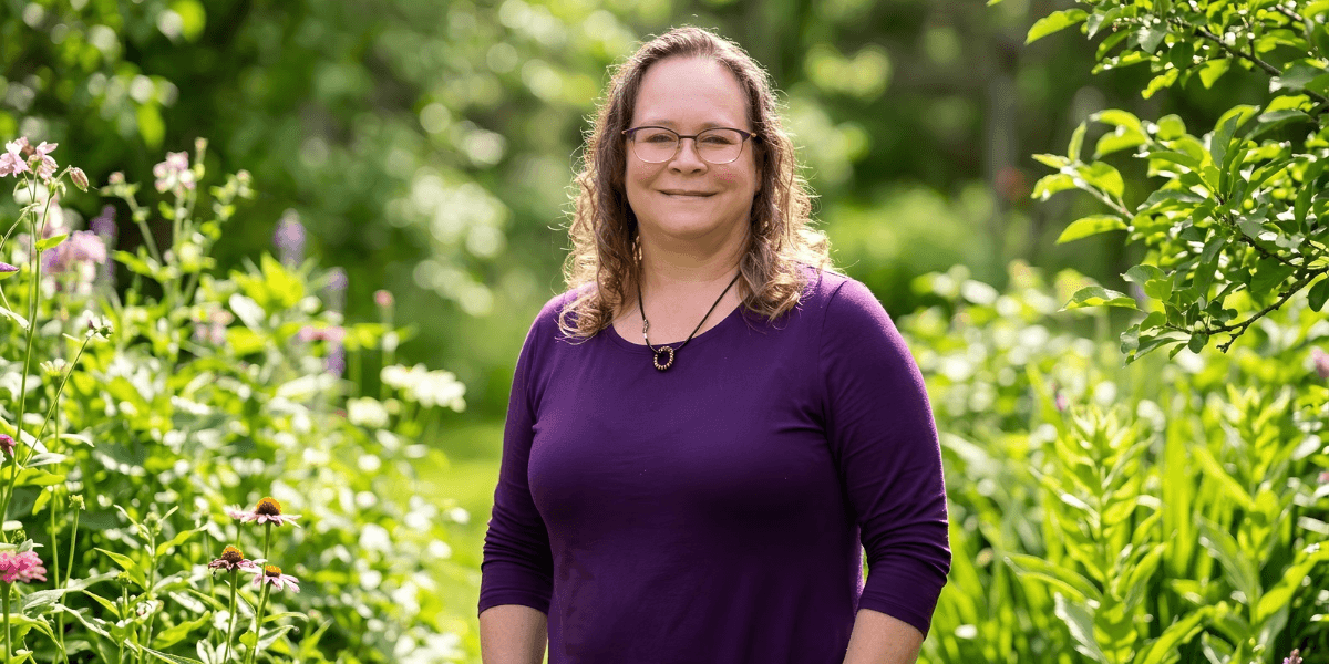 Smiling woman in purple top standing among colorful flowers in a lush garden