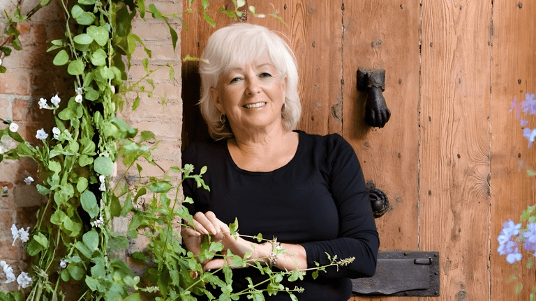 Patricia Sands smiling warmly, surrounded by greenery, leaning against a rustic wooden door.
