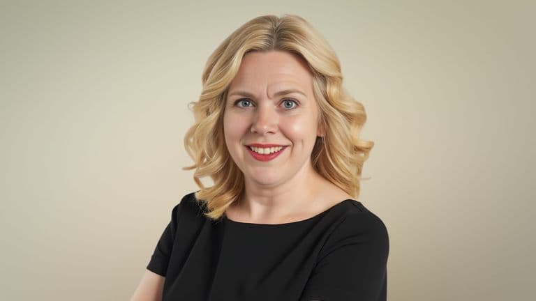 a smiling woman with blonde hair in a black top, standing against a neutral beige background.