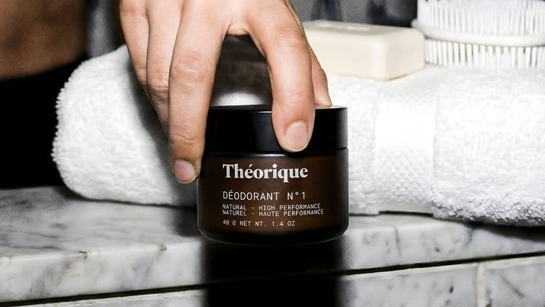 A hand holds Théorique Deodorant N°1 by a rolled white towel, soap, and brush on a marble surface, evoking a clean, minimalist feel.