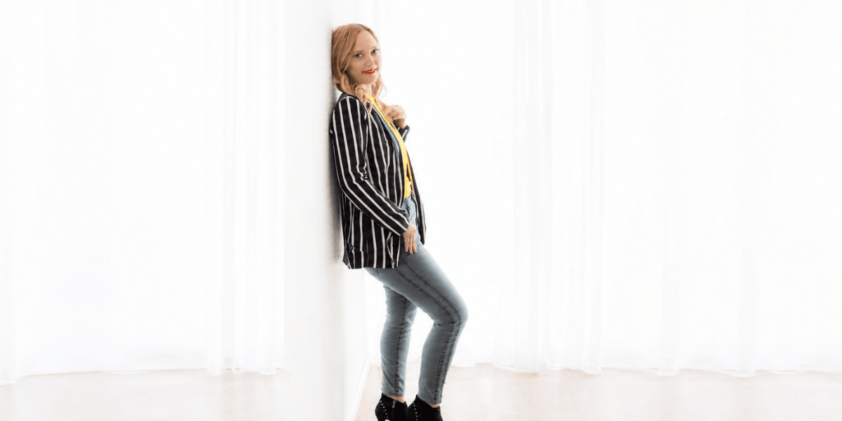 A woman in a striped blazer and jeans leans against a white wall