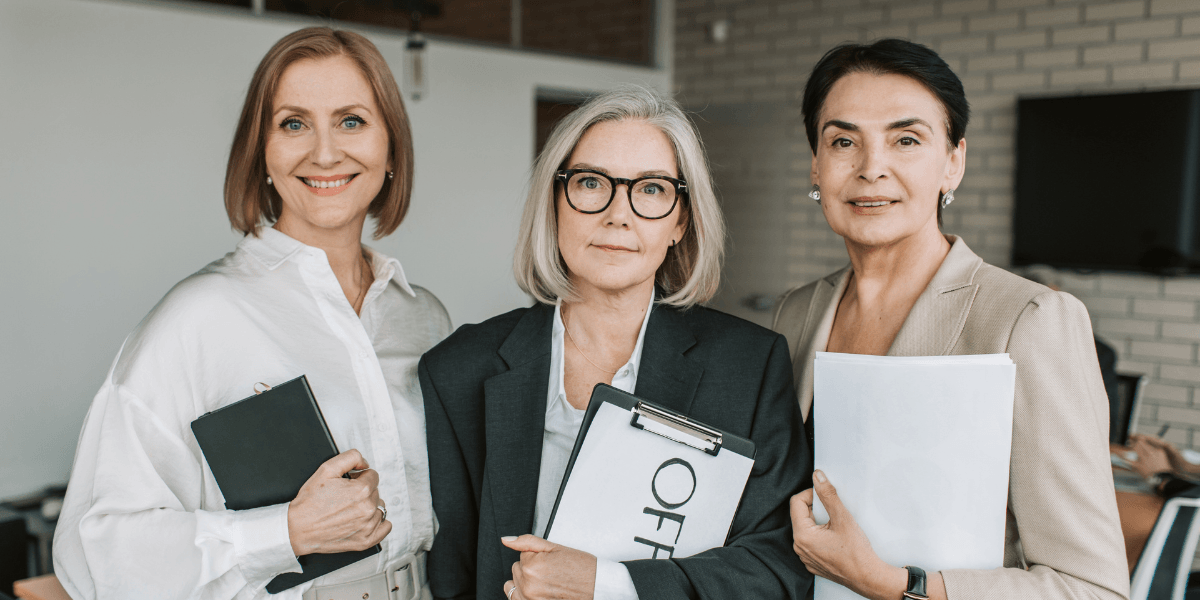 Three professional women stand together, holding clipboards and documents in an office setting