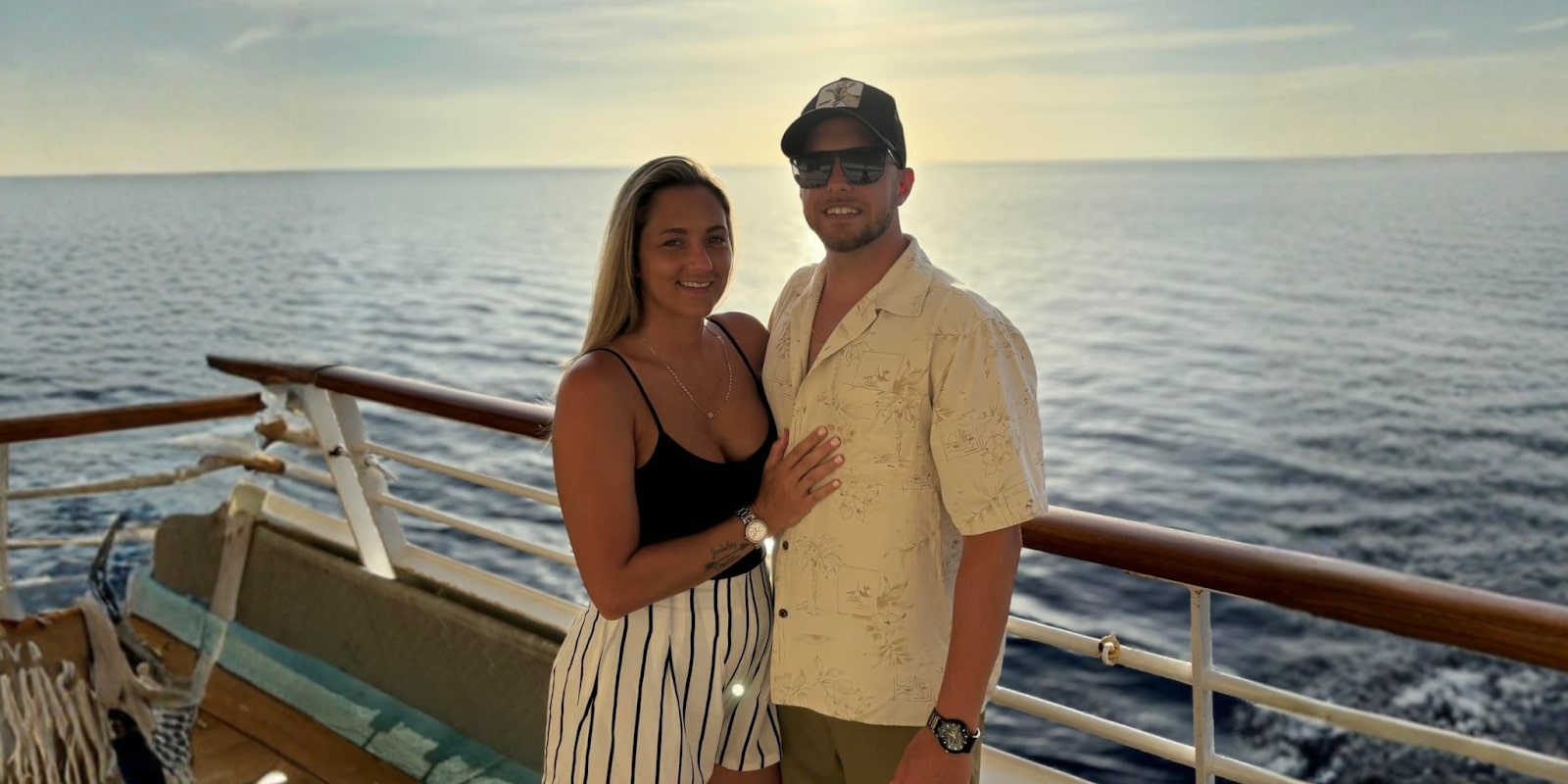 A couple stands together on a cruise ship deck, overlooking the ocean, smiling with a warm sunset in the background.