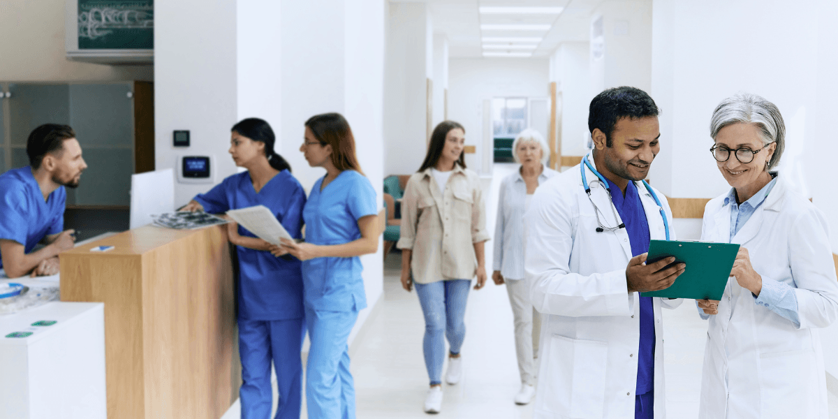 Doctors review a clipboard while nurses and patients stand in a hospital corridor.