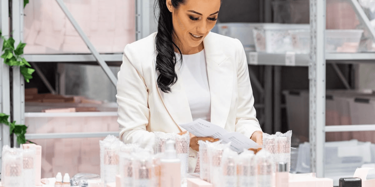 Woman in a white blazer reviewing paperwork amid beauty product packaging on a table
