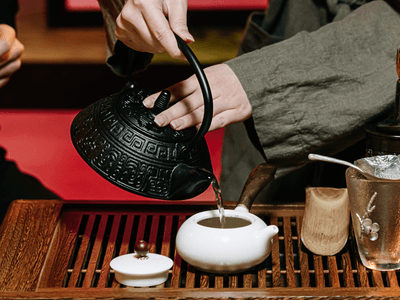 Person pouring tea from a traditional kettle into a small white teapot