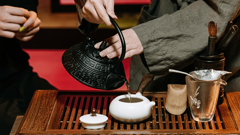 Person pouring tea from a traditional kettle into a small white teapot