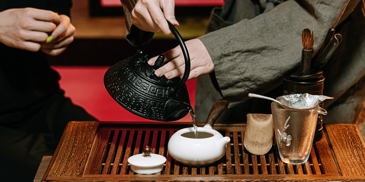 Person pouring tea from a traditional kettle into a small white teapot