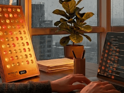 A person typing on a laptop beside a red light therapy device and a plant.