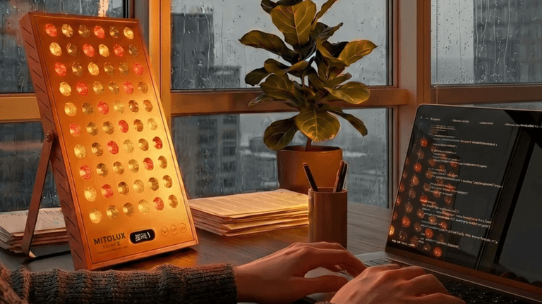 A person typing on a laptop beside a red light therapy device and a plant.