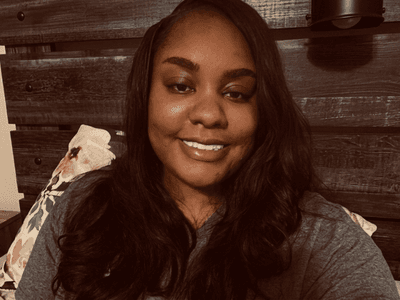 A woman with long hair smiles in a cozy bedroom setting.
