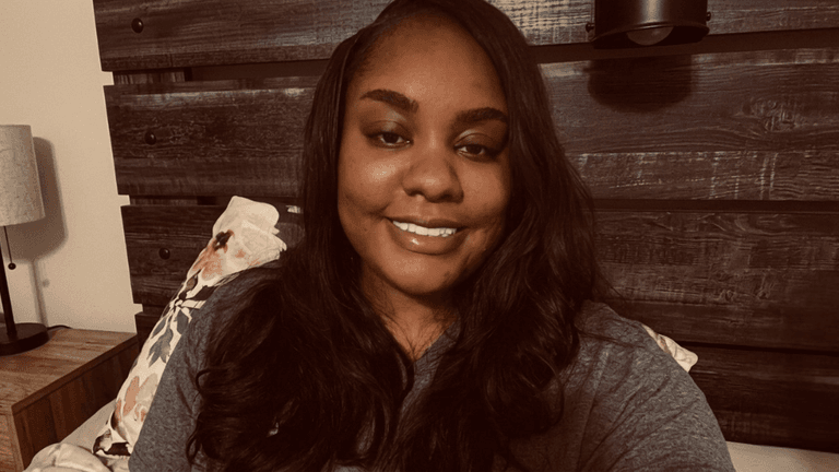 A woman with long hair smiles in a cozy bedroom setting.