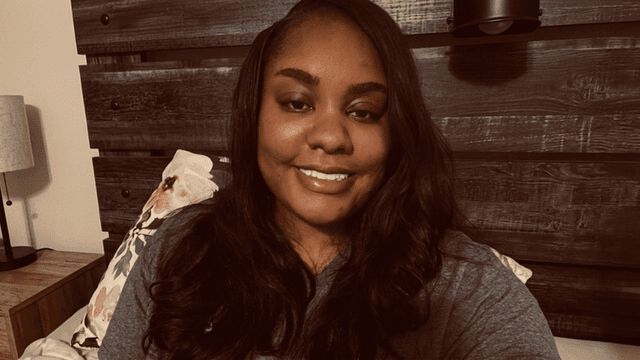 A woman with long hair smiles in a cozy bedroom setting.