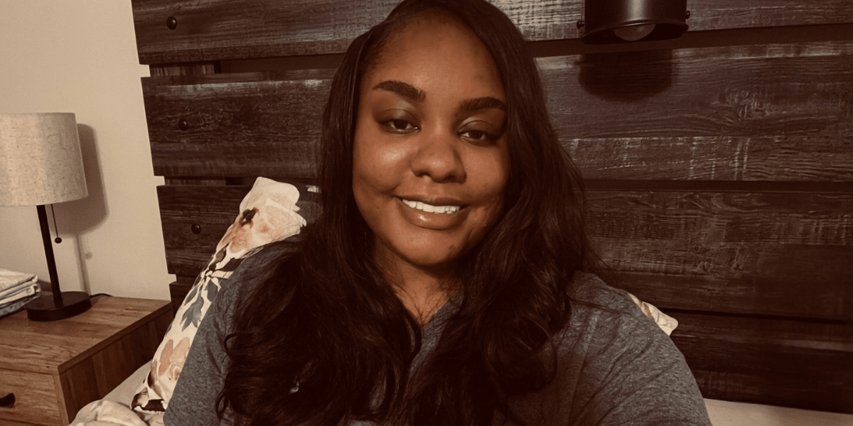 A woman with long hair smiles in a cozy bedroom setting.