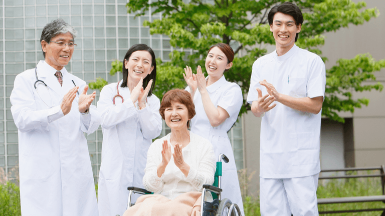 A smiling elderly woman in a wheelchair is cheered on by four medical staff members