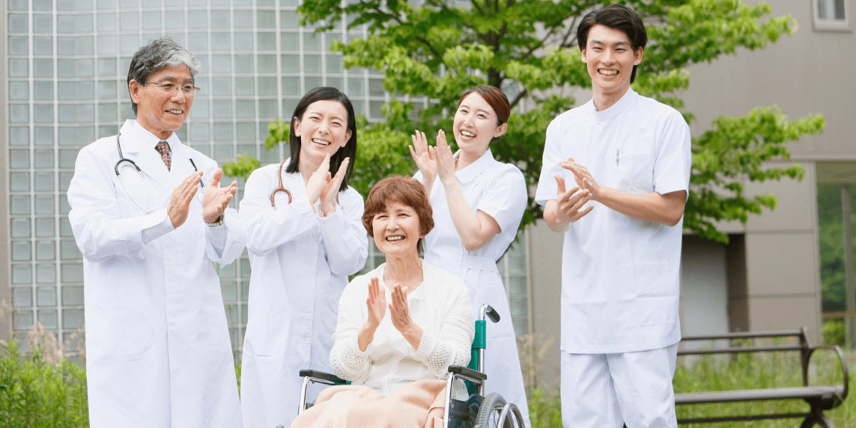 A smiling elderly woman in a wheelchair is cheered on by four medical staff members