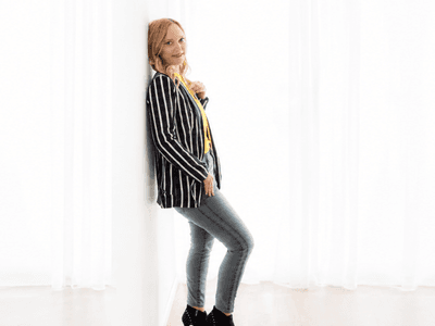 A woman in a striped blazer and jeans leans against a white wall