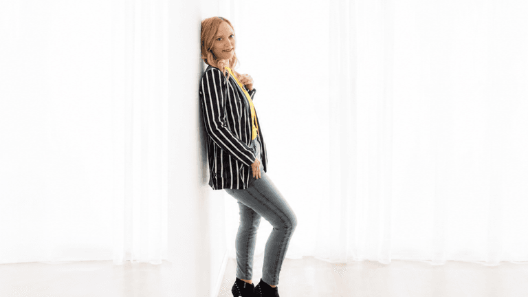 A woman in a striped blazer and jeans leans against a white wall
