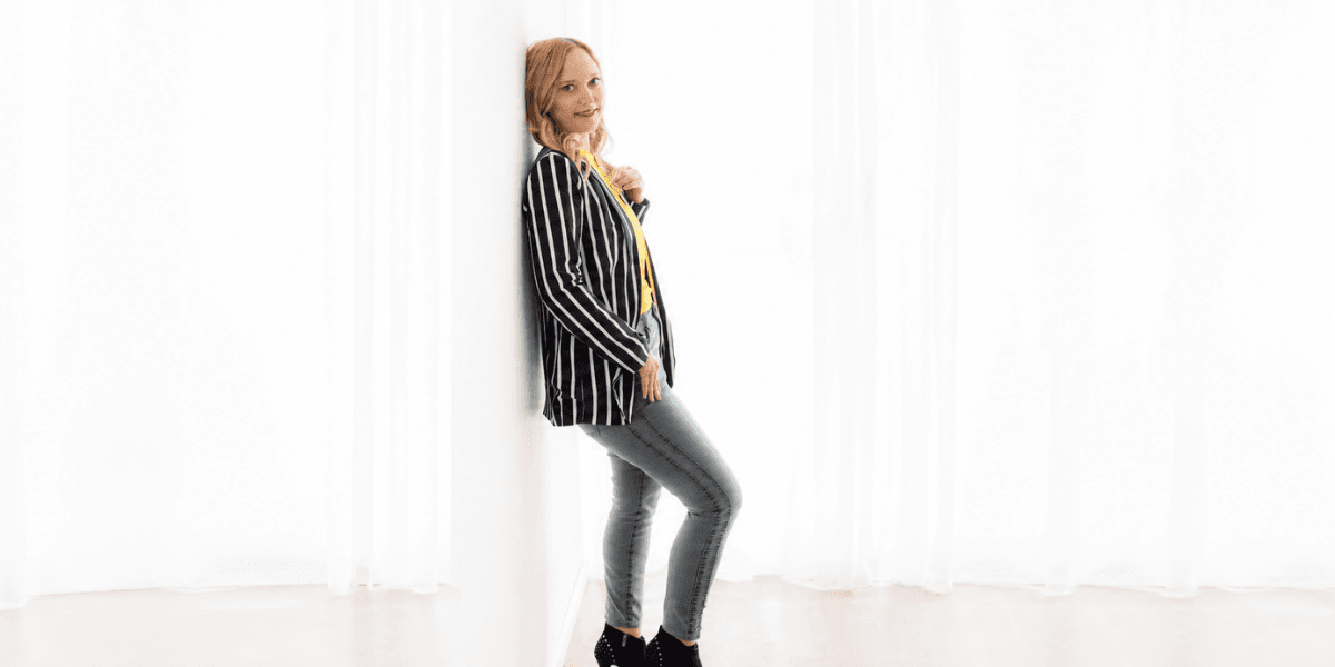 A woman in a striped blazer and jeans leans against a white wall