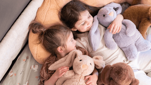 Two young girls cuddle with plush toys on a cozy bed.
