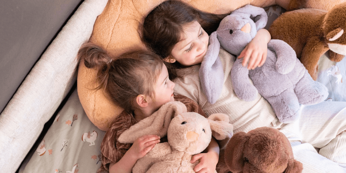 Two young girls cuddle with plush toys on a cozy bed.