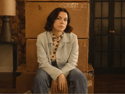 A woman sits on stacked boxes in a vintage room with floral wallpaper