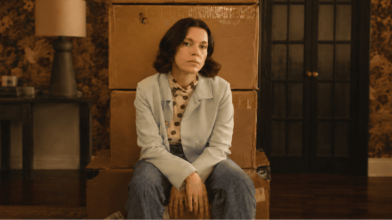 A woman sits on stacked boxes in a vintage room with floral wallpaper
