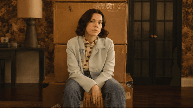 A woman sits on stacked boxes in a vintage room with floral wallpaper