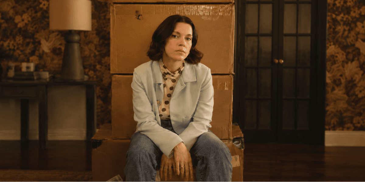 A woman sits on stacked boxes in a vintage room with floral wallpaper