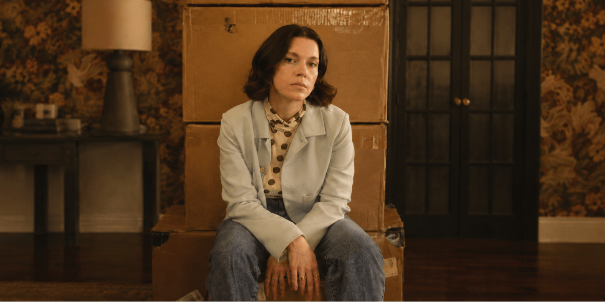 A woman sits on stacked boxes in a vintage room with floral wallpaper