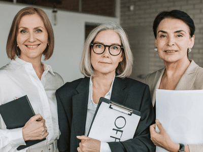 Three professional women stand together, holding clipboards and documents in an office setting