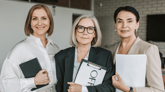 Three professional women stand together, holding clipboards and documents in an office setting