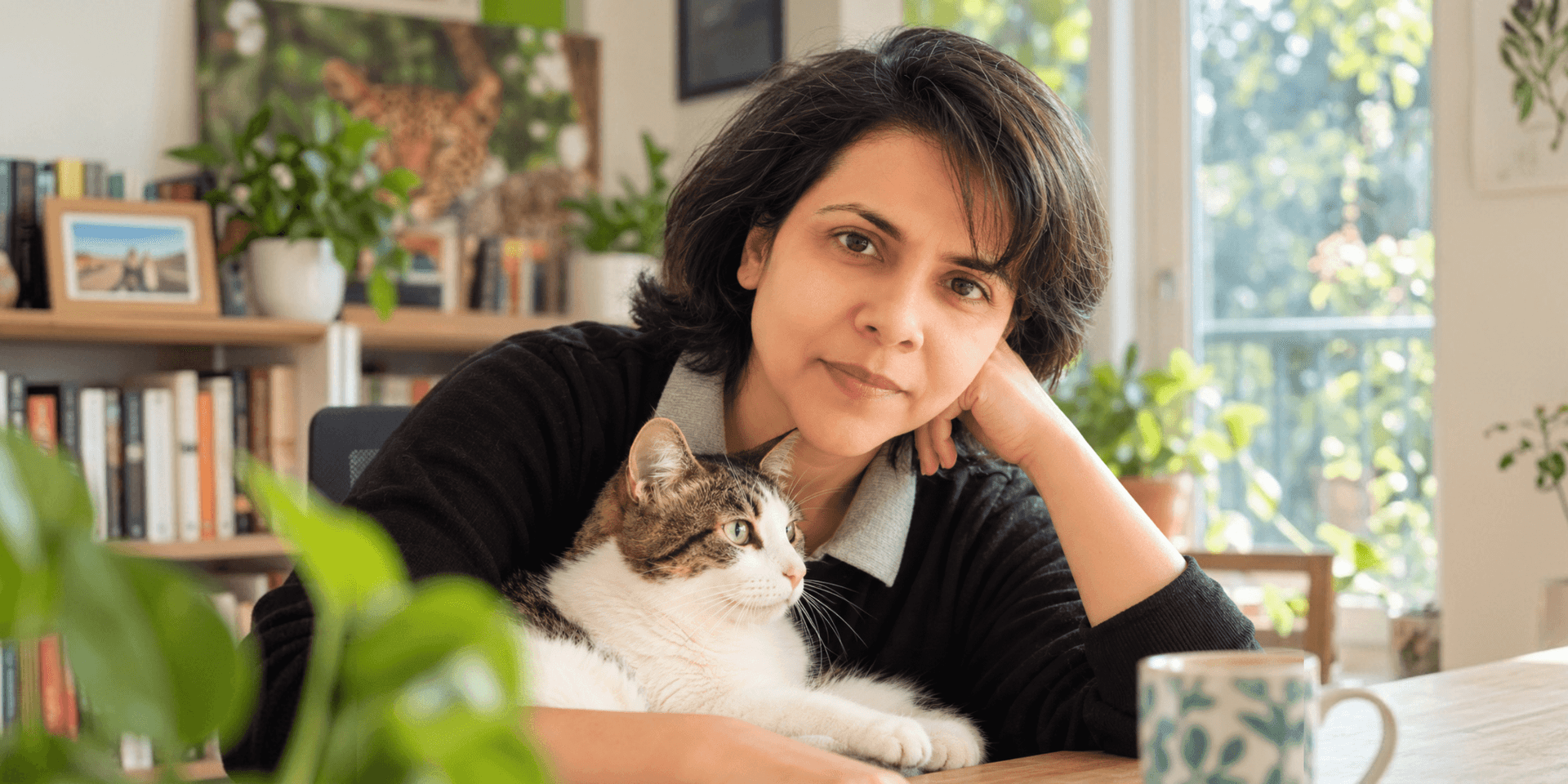 A woman with dark hair sits at a table with a cat and plants nearby