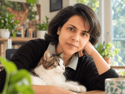 A woman with dark hair sits at a table with a cat and plants nearby