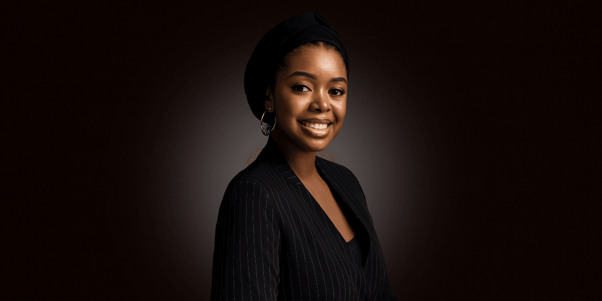 A smiling woman in a black pinstripe suit and headscarf against a dark background