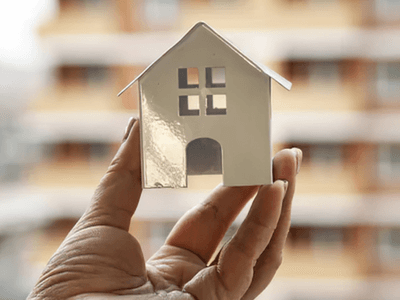 A hand holds a small white model house against a blurred building background