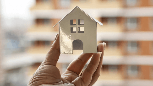 A hand holds a small white model house against a blurred building background
