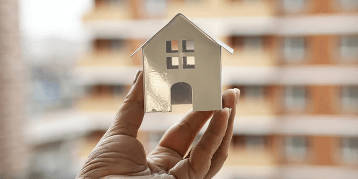 A hand holds a small white model house against a blurred building background
