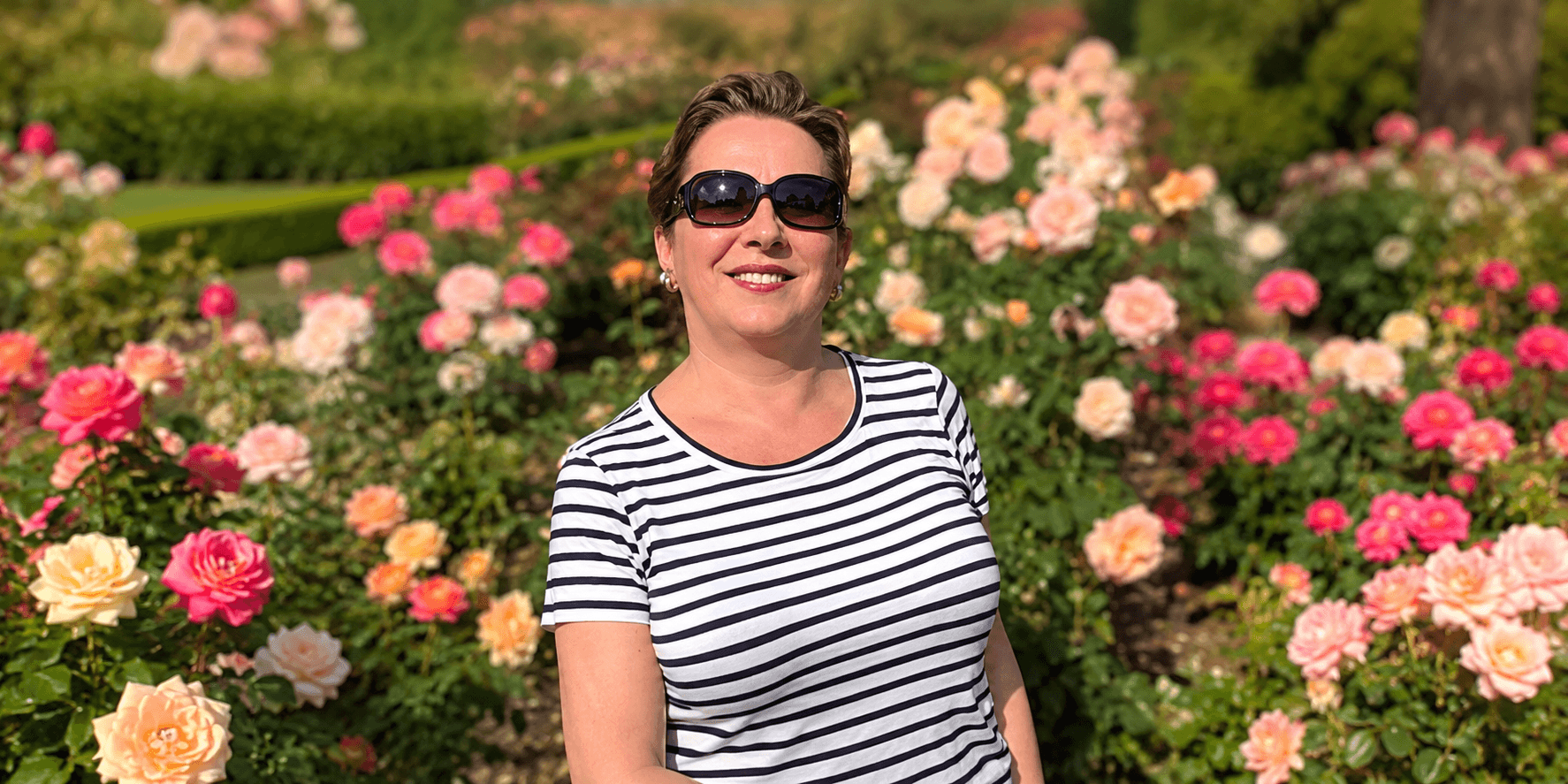 Woman in sunglasses poses among blooming roses in a vibrant garden setting