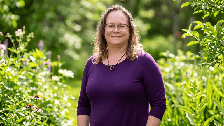 Smiling woman in purple top standing among colorful flowers in a lush garden