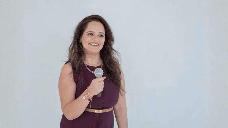 A woman with long hair smiles while holding a microphone against a light background