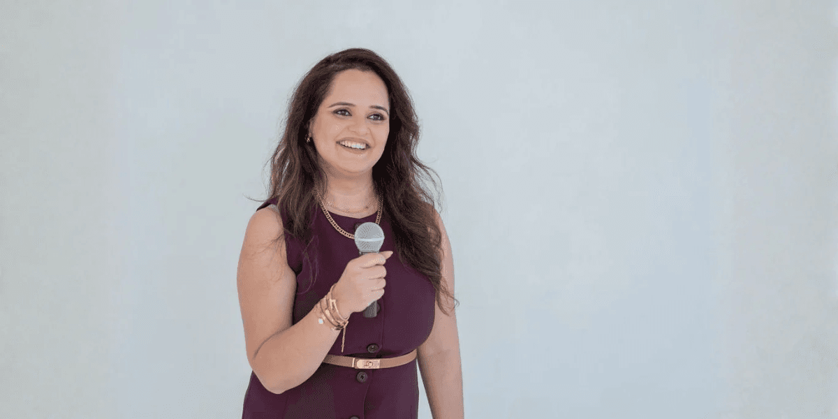 A woman with long hair smiles while holding a microphone against a light background