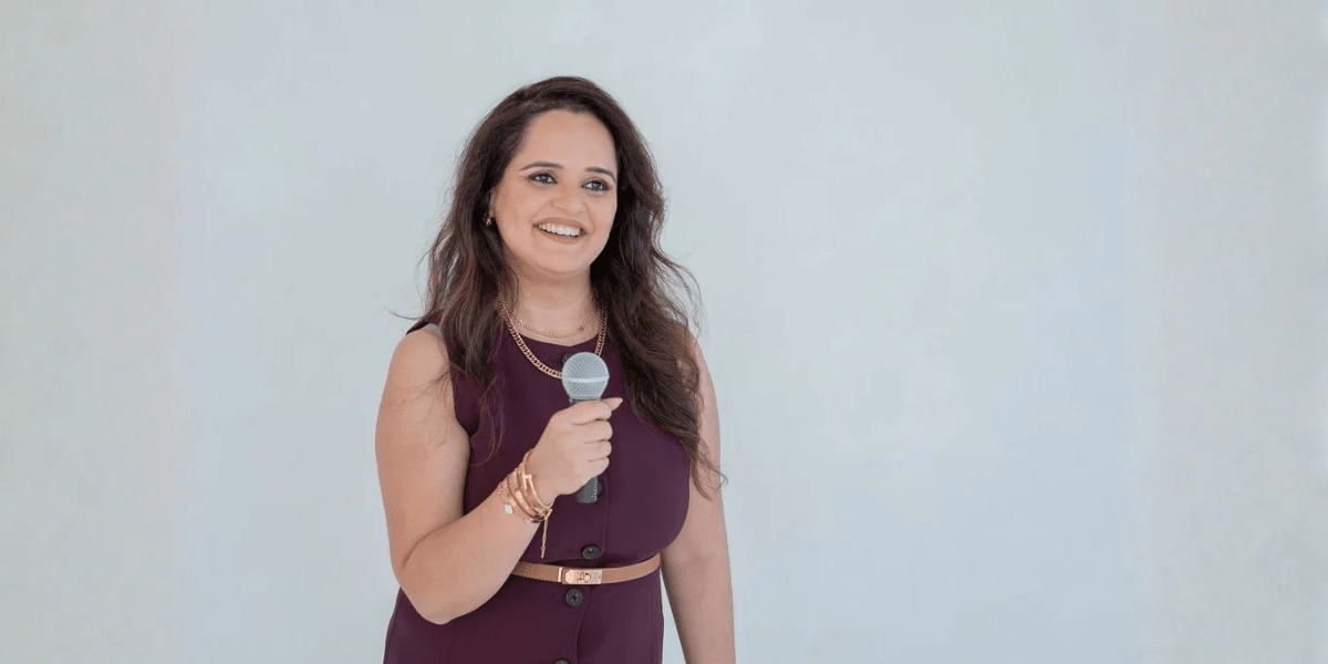 A woman with long hair smiles while holding a microphone against a light background