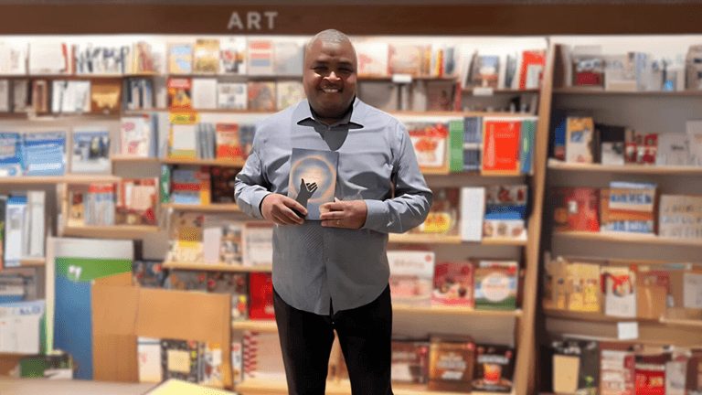 Kevin Howard holds a copy of his book Onward, At Last in front of a colorful bookshelf, standing in a bookstore with a warm smile.