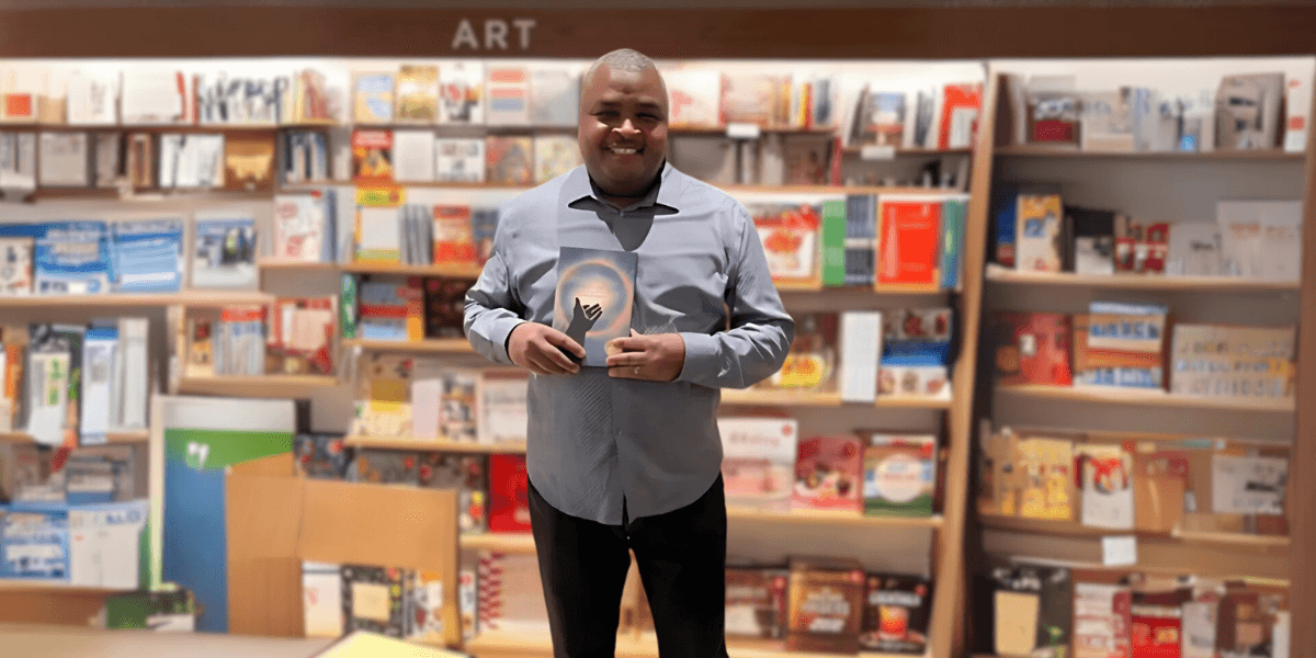 Kevin Howard holds a copy of his book Onward, At Last in front of a colorful bookshelf, standing in a bookstore with a warm smile.