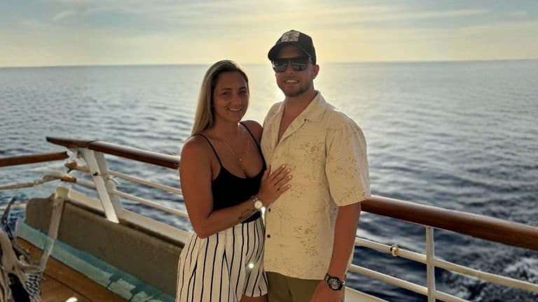 A couple stands together on a cruise ship deck, overlooking the ocean, smiling with a warm sunset in the background.