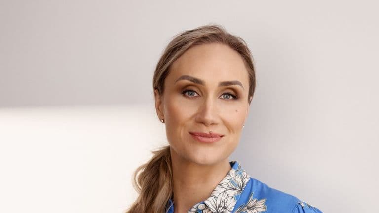 Simona Smaidžiūnaitė with a confident smile, wearing a blue blouse with floral patterns, styled with soft makeup and ponytail.