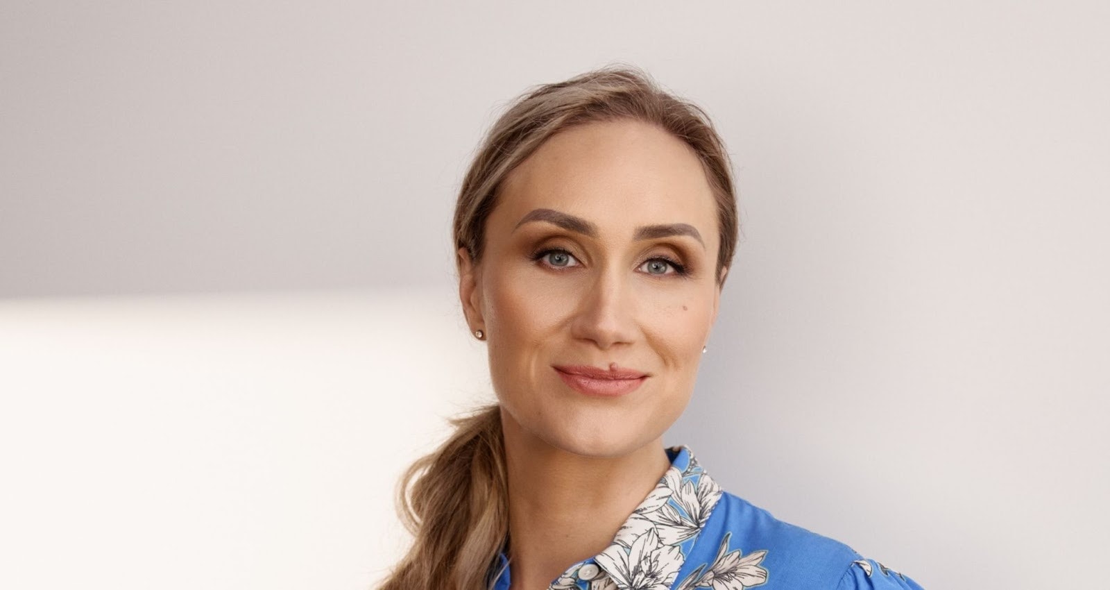 Simona Smaidžiūnaitė with a confident smile, wearing a blue blouse with floral patterns, styled with soft makeup and ponytail.