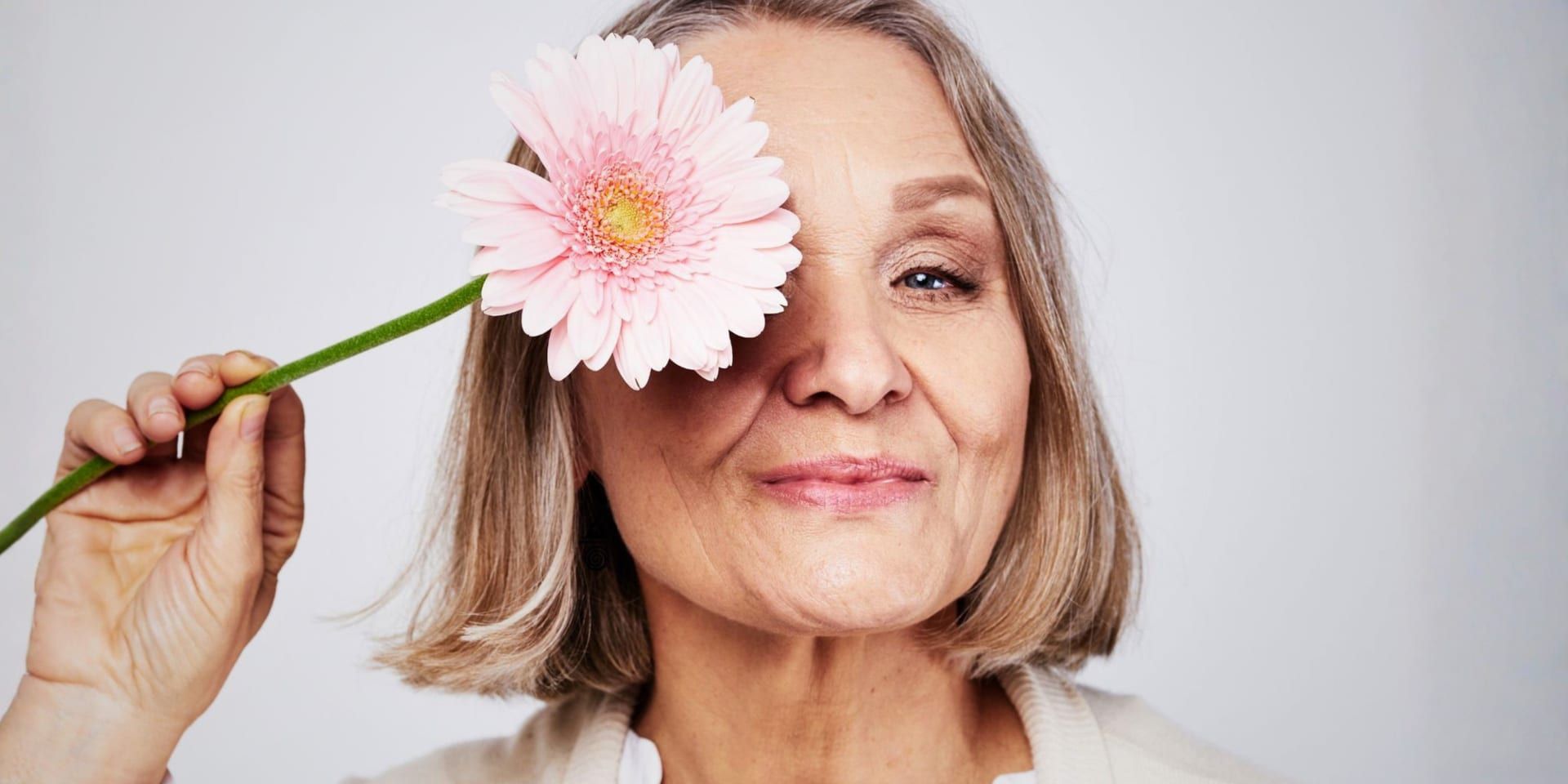 Older woman smiling while holding a pink flower over one eye against a soft neutral background.